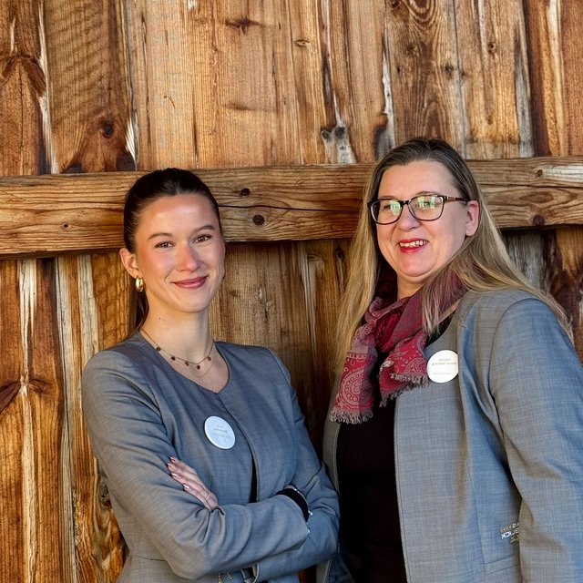 Lerch Businesswelten - Inspirationen an einem Tag Zwei Frauen in grauen Blazern vor einer Holzwand lächeln in die Kamera