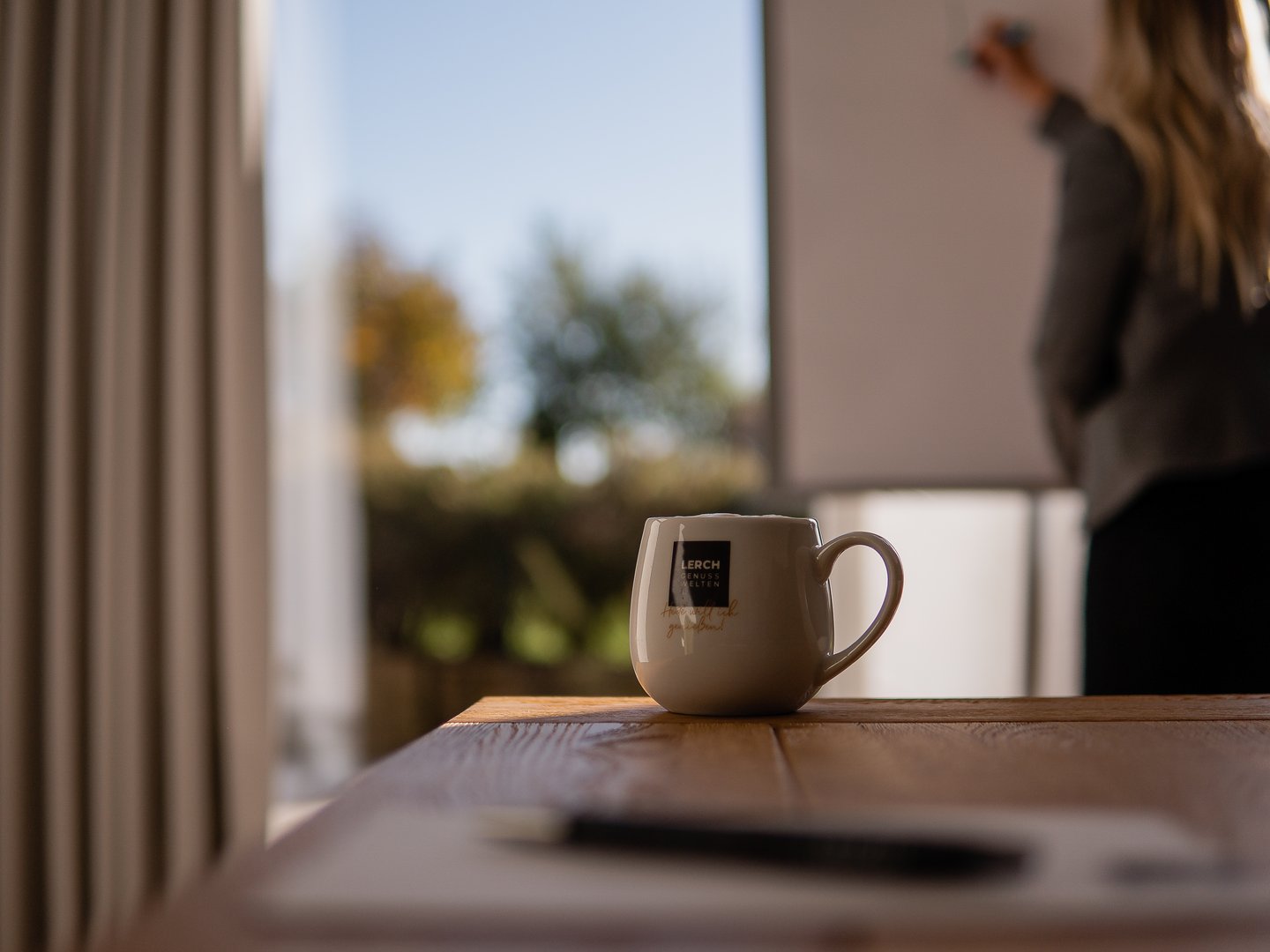 Lerch Businesswelten - Inspirationen an einem Tag Kaffeetasse auf Holztisch mit verschwommener Person vor Whiteboard