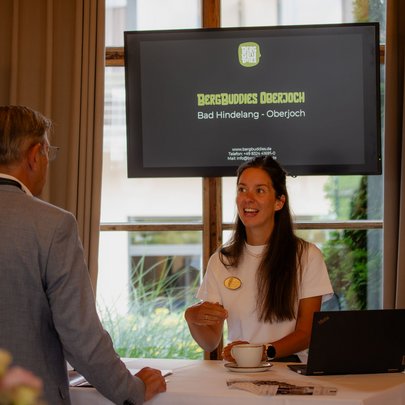 Lerch Businesswelten - Inspirationen an einem Tag Frau spricht an einem Tisch mit Mann vor Bildschirm von BergBuddies Oberjoch
